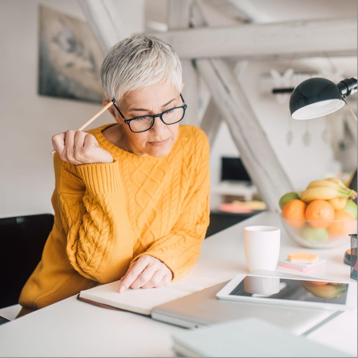 A thoughtful older woman in a yellow sweater tapping a pencil against her head while reading at her desk