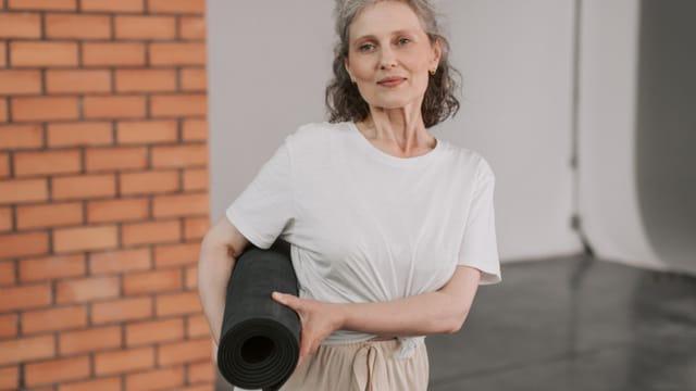 A smiling older woman holding a rolled-up exercise mat under her arm