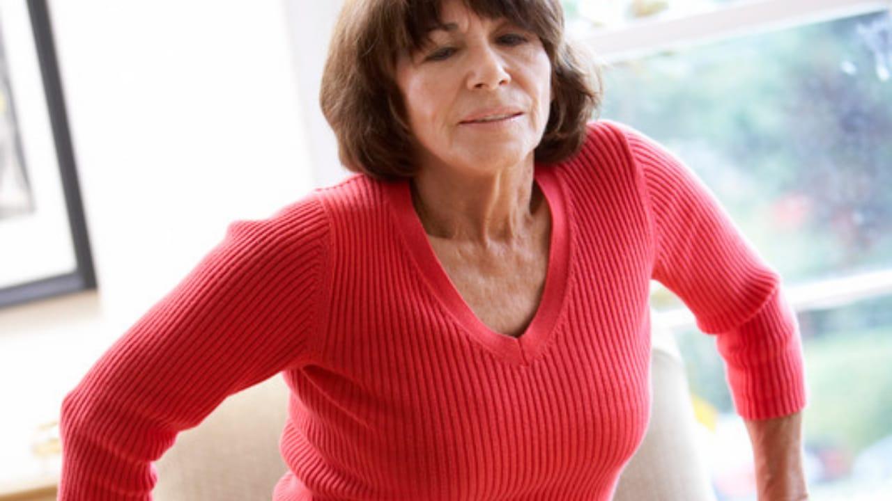 An older woman in a red sweater pushing herself up from an armchair with both hands