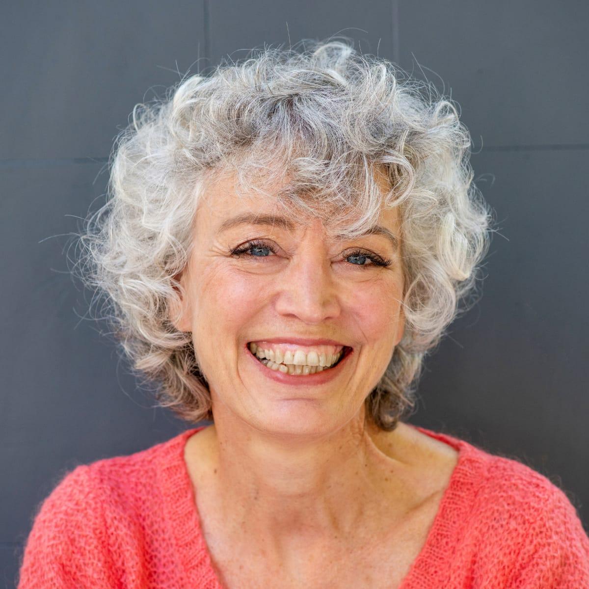 A smiling older woman with curly gray hair in a coral sweater