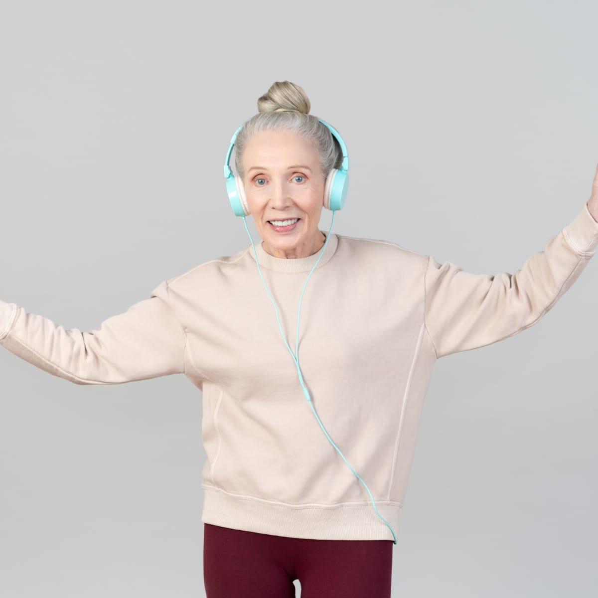 A smiling older woman in headphones dancing with her arms out