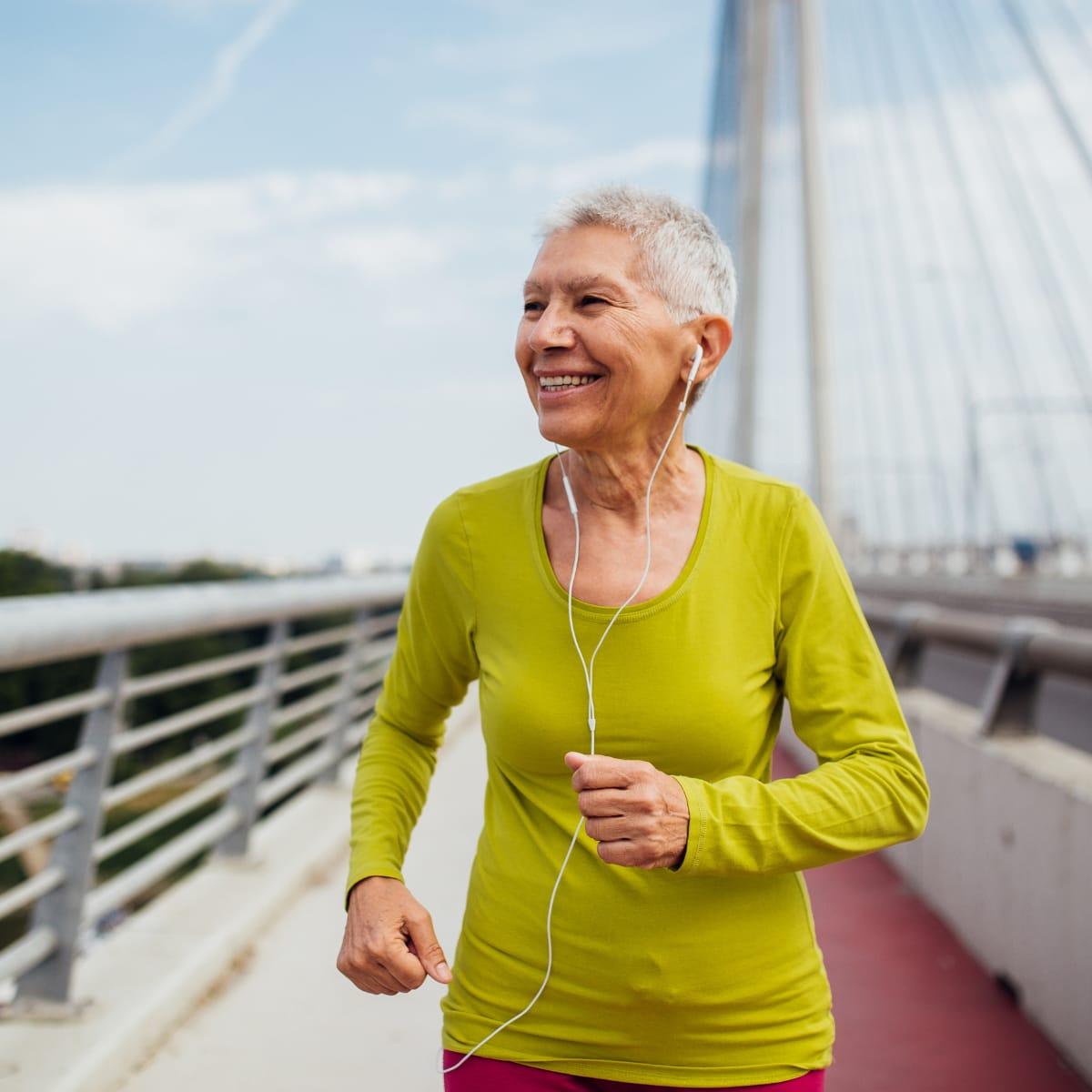 A smiling older woman jogging across a bridge with earbuds in