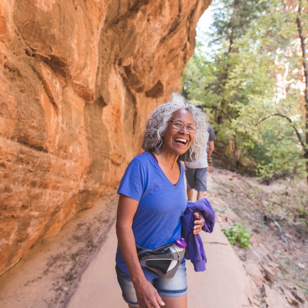 A gray-haired woman in a blue T-shirt laughing on a canyon trail beside a red rock wall