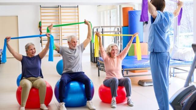Older adults sitting on exercise balls, stretching resistance bands overhead with an instructor