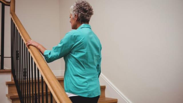 An older woman in a teal shirt starting up a staircase at home, holding the wooden handrail