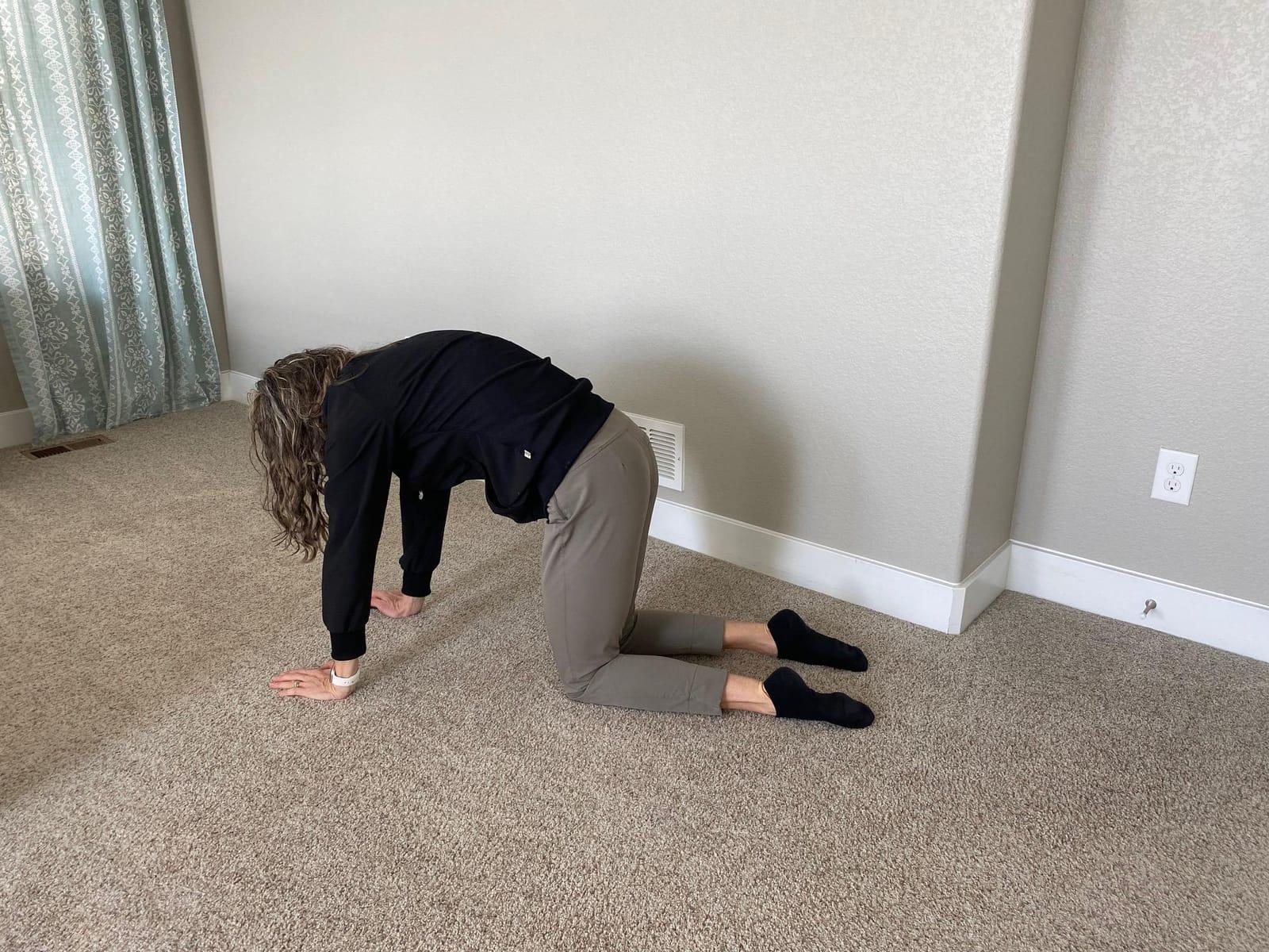 A woman on her hands and knees on a carpeted floor, rounding her back in the cat position
