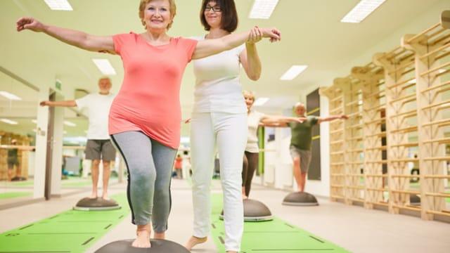 An older woman balancing on a half-ball trainer with arms out, next to an instructor in a bright gym