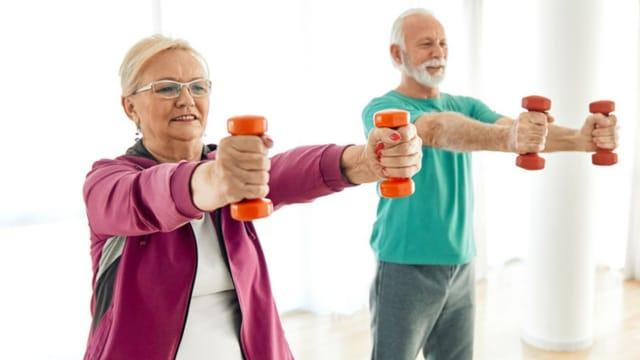 An older woman and man holding small orange dumbbells straight out in front of them
