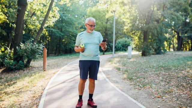 An older man in a light blue T-shirt walking on a tree-lined park path, looking at his phone
