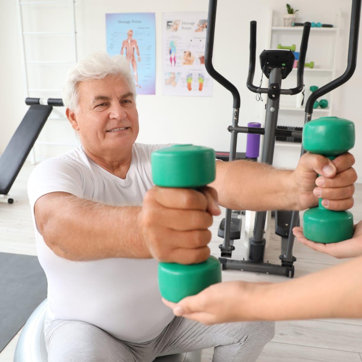 An older man seated on an exercise ball pressing two green dumbbells forward as a trainer supports his wrists