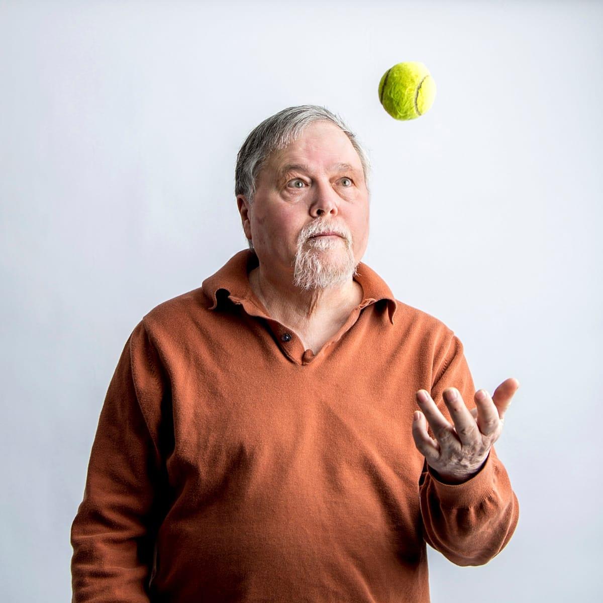 An older man with a white beard tossing a tennis ball and watching it in the air