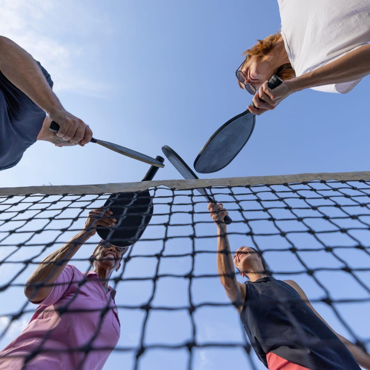 Low-angle view of smiling players raising their pickleball paddles together over the net against a blue sky