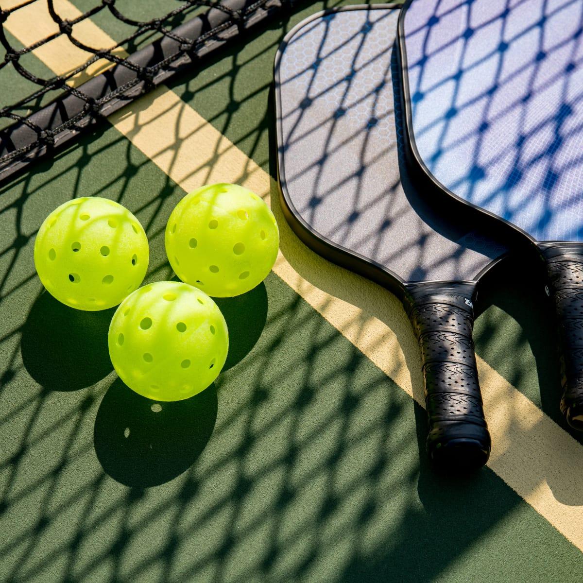 Three yellow-green pickleballs and two paddles lying on a court in the shadow of the net