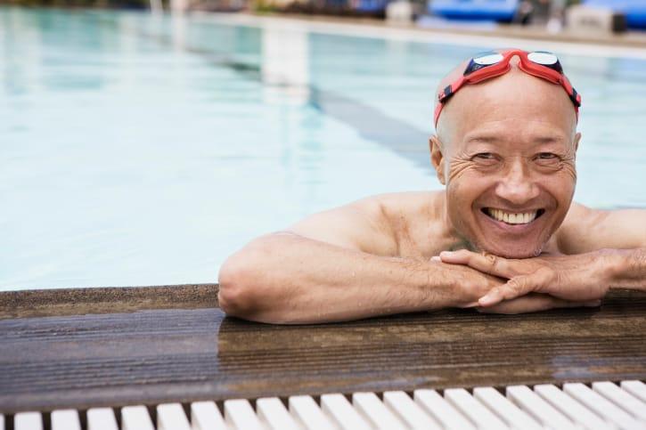 A grinning older man in red swim goggles resting his arms on the edge of a pool