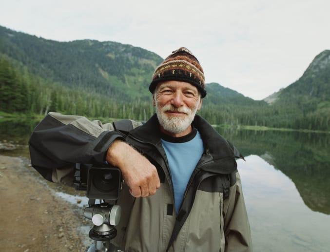 A smiling older man in a knit hat leaning on a camera tripod beside a mountain lake