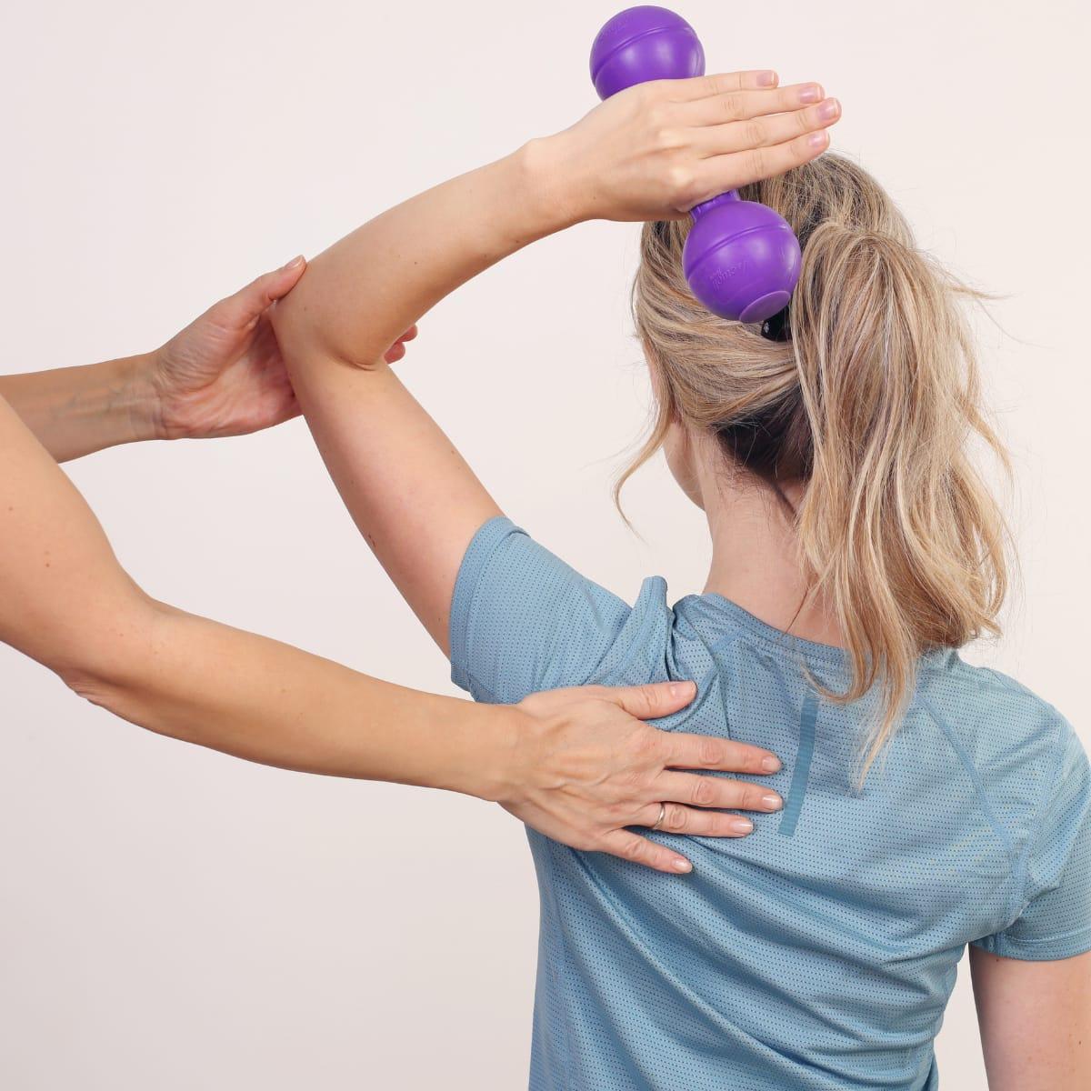 A trainer guiding a woman's shoulder blade as she lifts a small purple dumbbell overhead