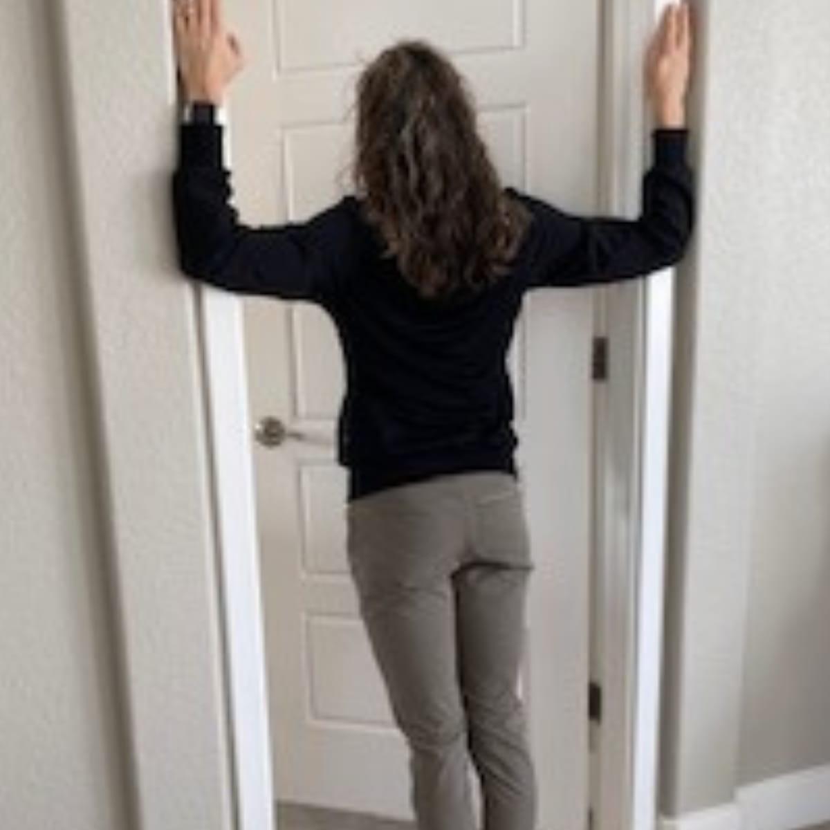 A woman standing in an open doorway with her hands on either side of the frame, leaning forward into a chest stretch