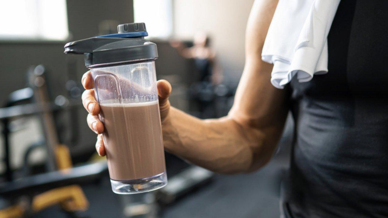 A hand holding a chocolate protein shake in a gym, with a towel over the shoulder
