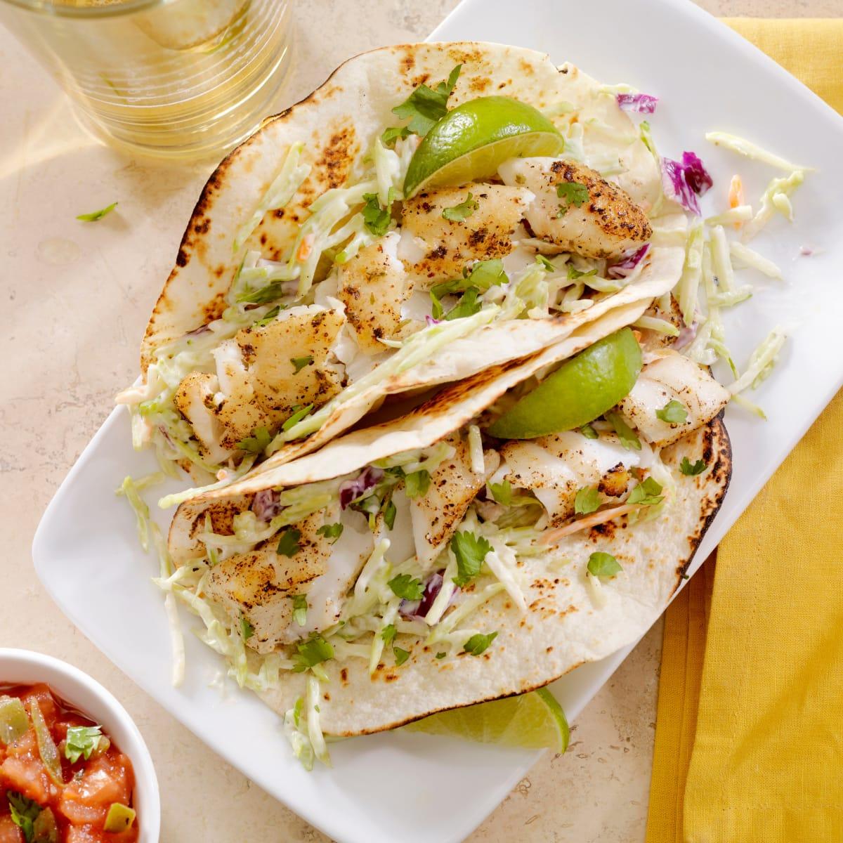 Two grilled fish tacos with shredded cabbage slaw, cilantro and lime wedges on a white plate