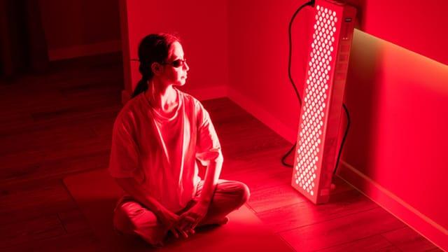 A woman in protective glasses sitting cross-legged on the floor beside a tall red light therapy panel
