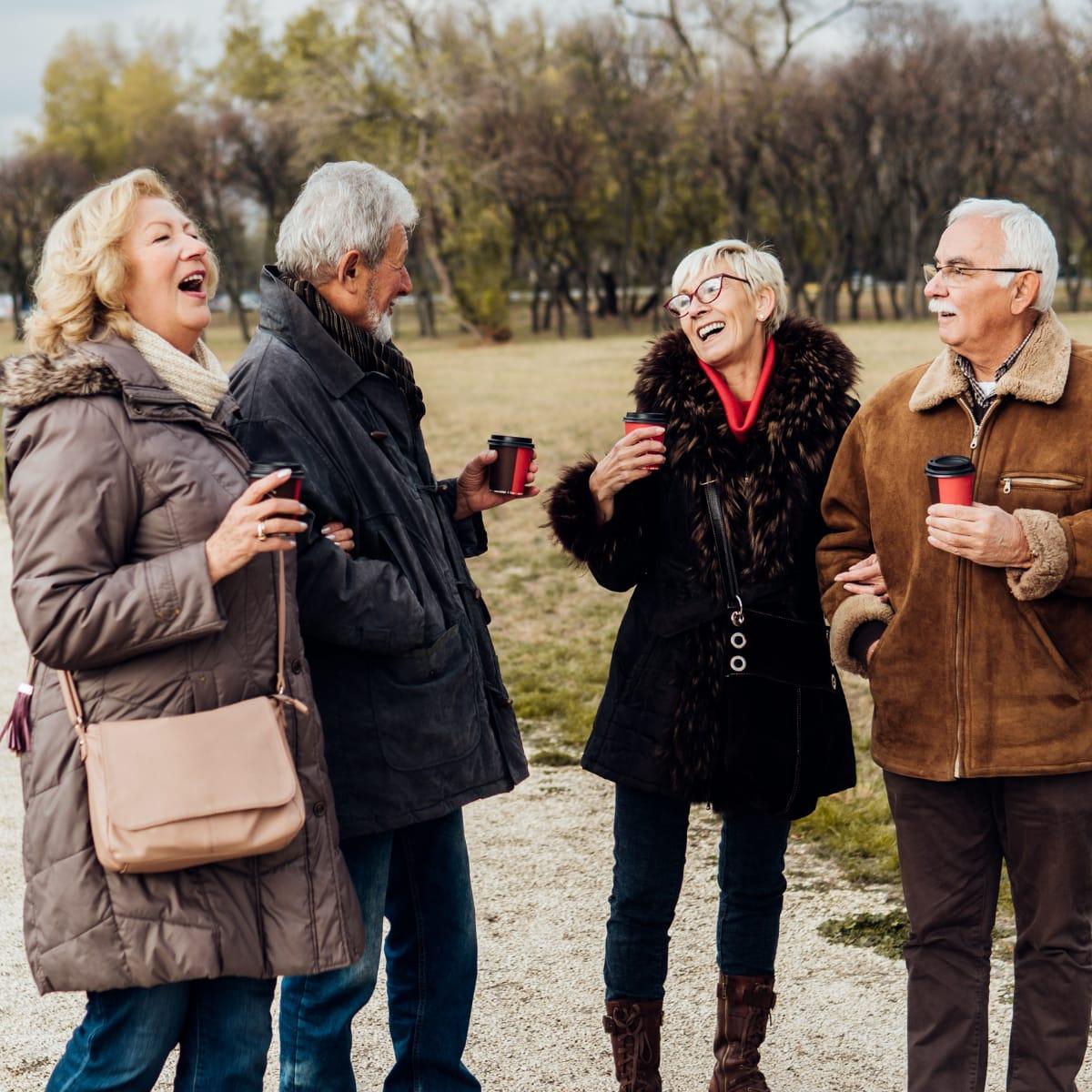 Four older friends laughing together outdoors in winter coats, holding coffee cups