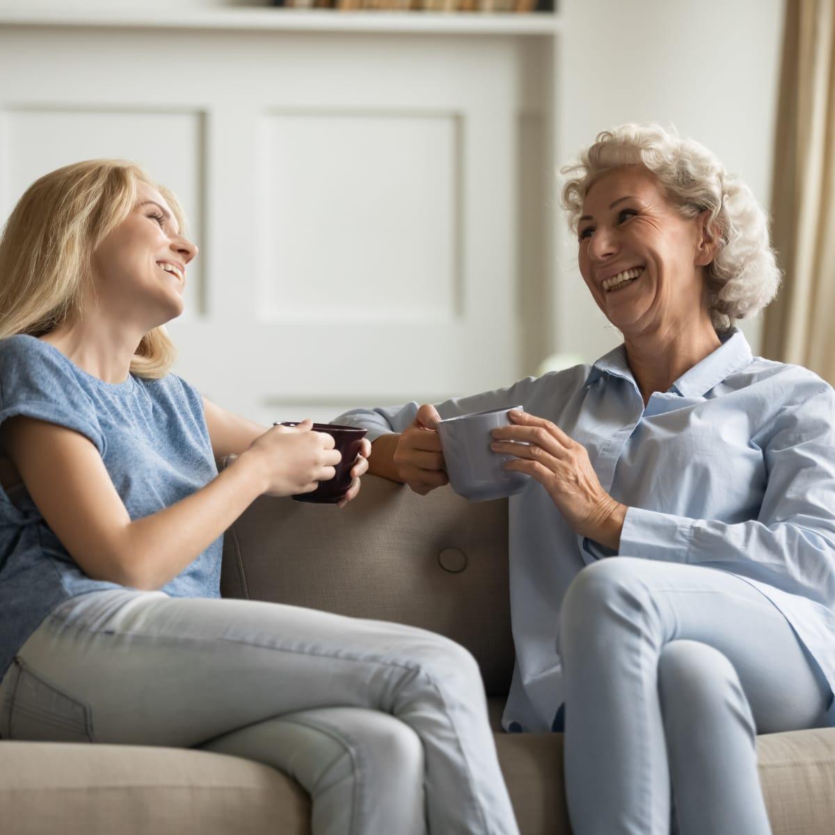 An older woman and a younger woman laughing together on a sofa with mugs in hand