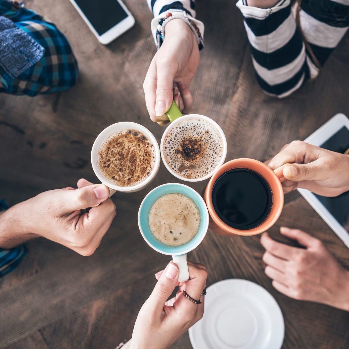 Four people clinking mugs of coffee together over a wooden table, seen from above