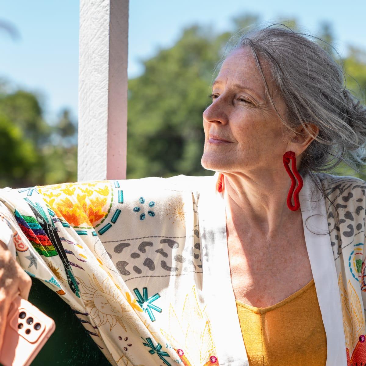 A smiling older woman with long gray hair and red earrings enjoying the morning sun outdoors