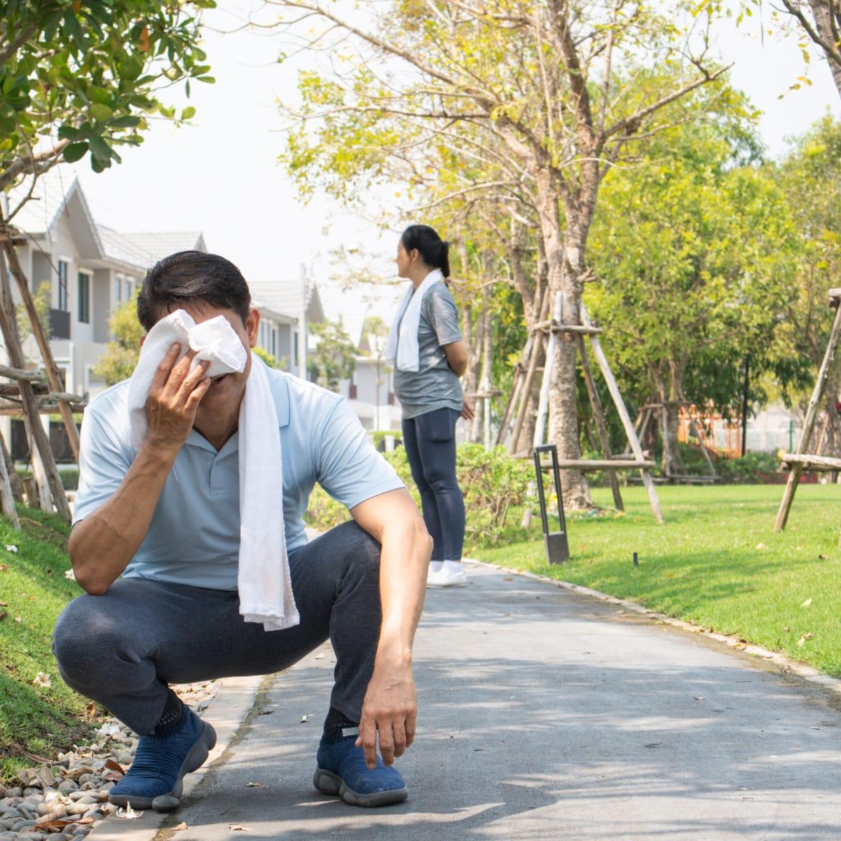 A man crouching on a park path wiping his face with a towel after a hard workout