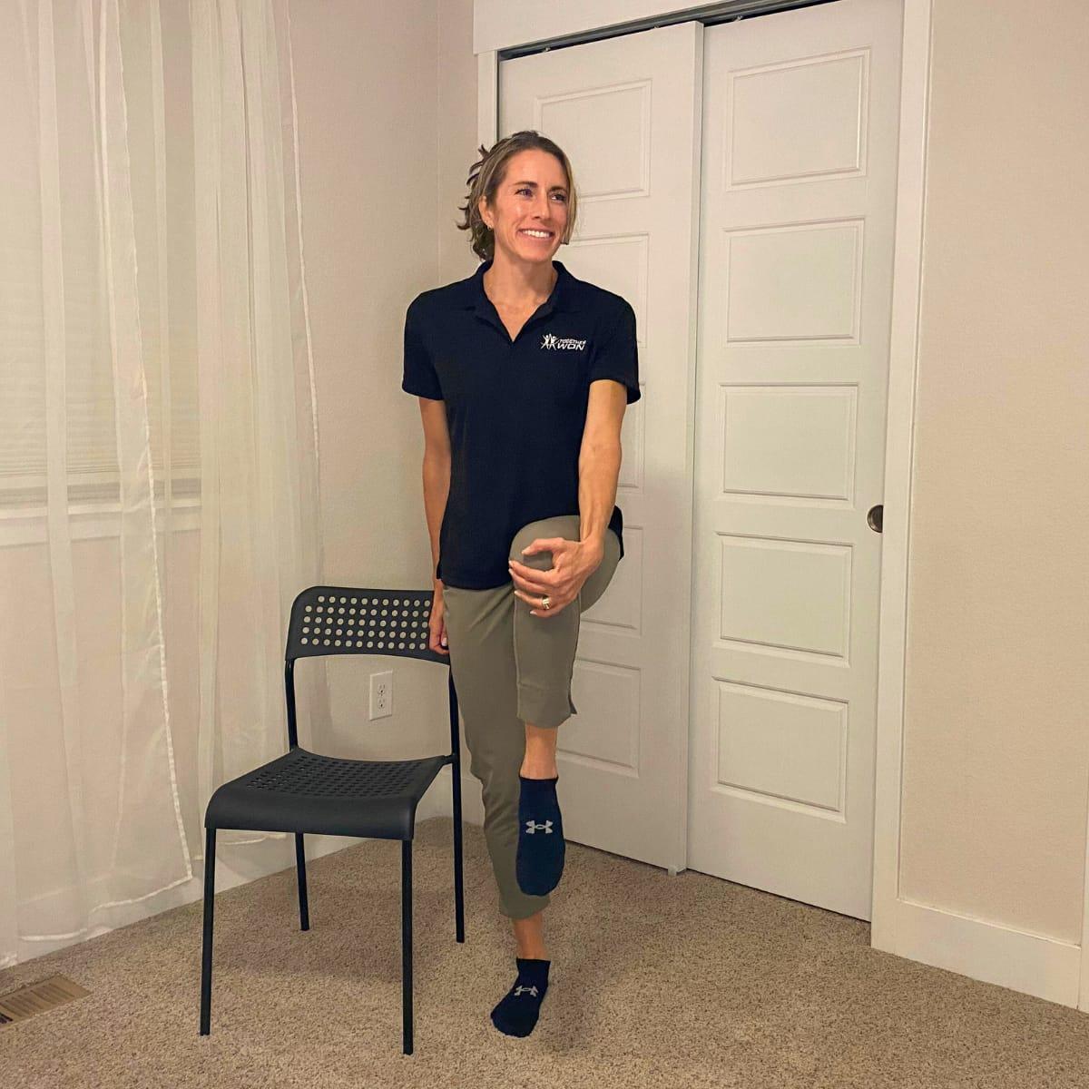 A trainer standing on one leg next to a chair, holding her raised knee with one hand