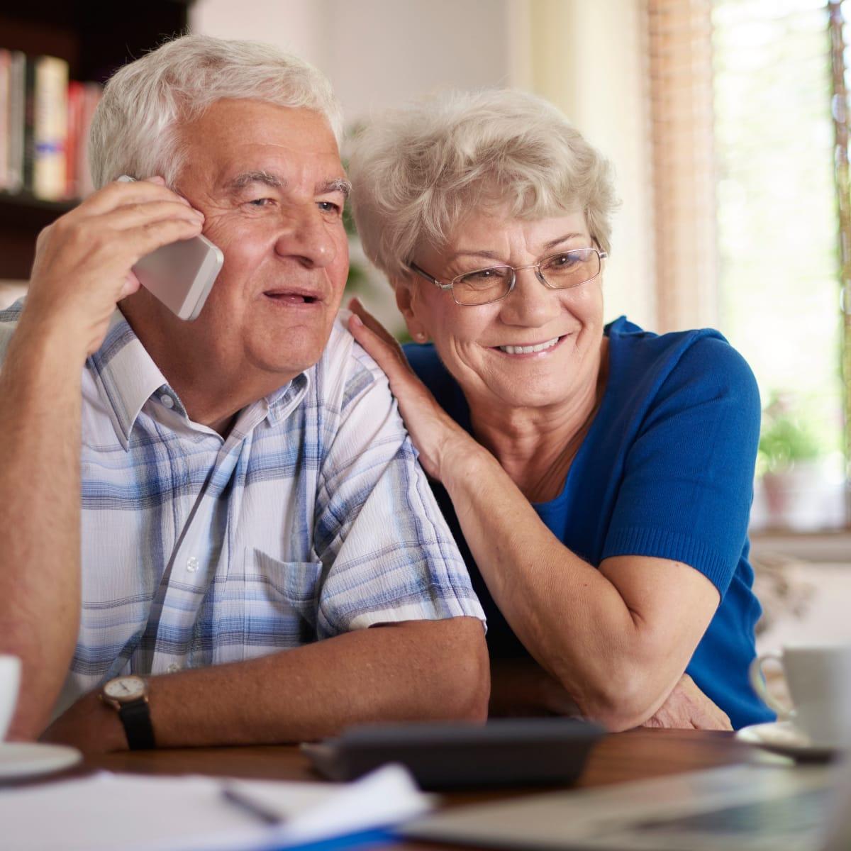 An older man chatting on the phone while his wife leans on his shoulder, smiling