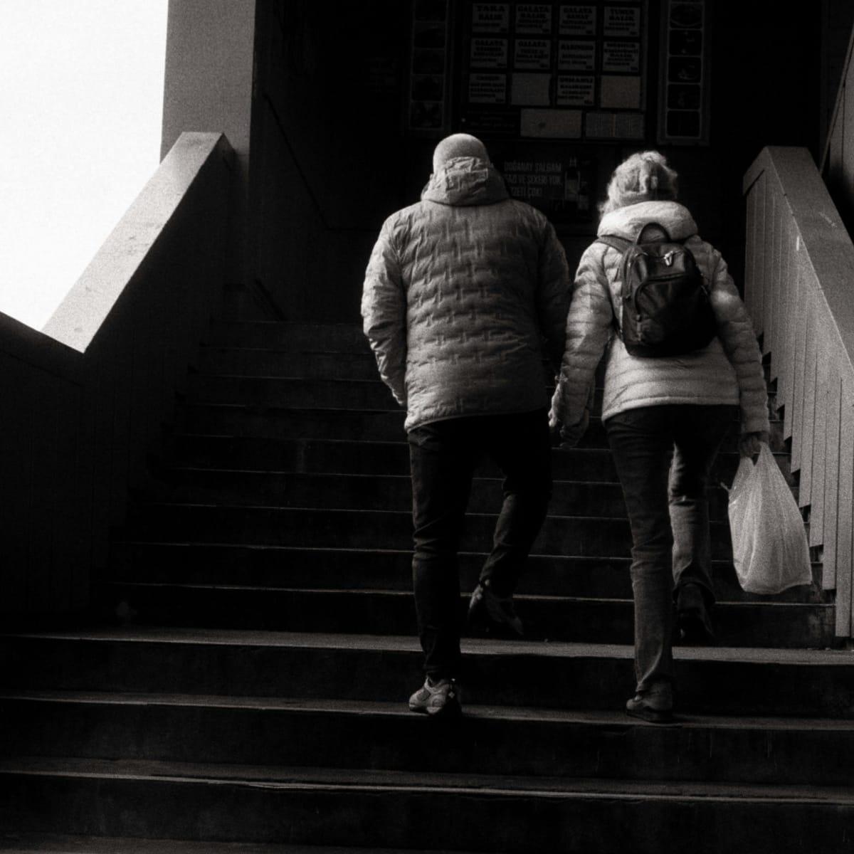 Black-and-white photo of an older couple in puffy jackets climbing an indoor staircase side by side