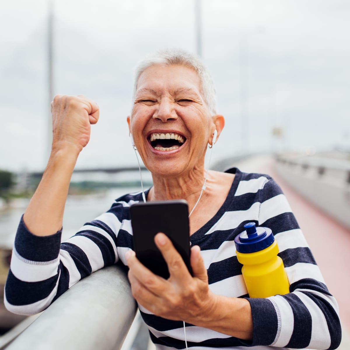 A laughing older woman in a striped top pumping her fist while holding a phone and water bottle outdoors