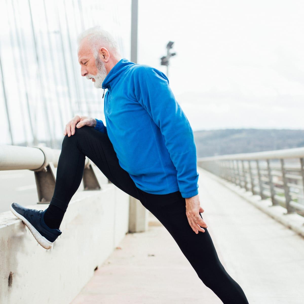 An older man in a blue fleece stretching his leg on a railing along a bridge walkway