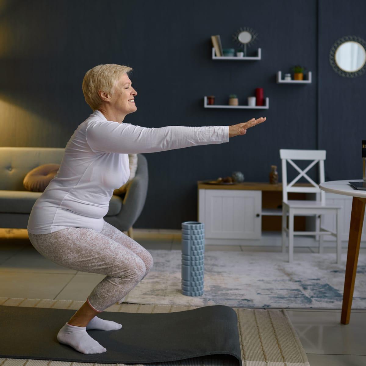 A smiling older woman doing a bodyweight squat with arms extended on a mat in her living room