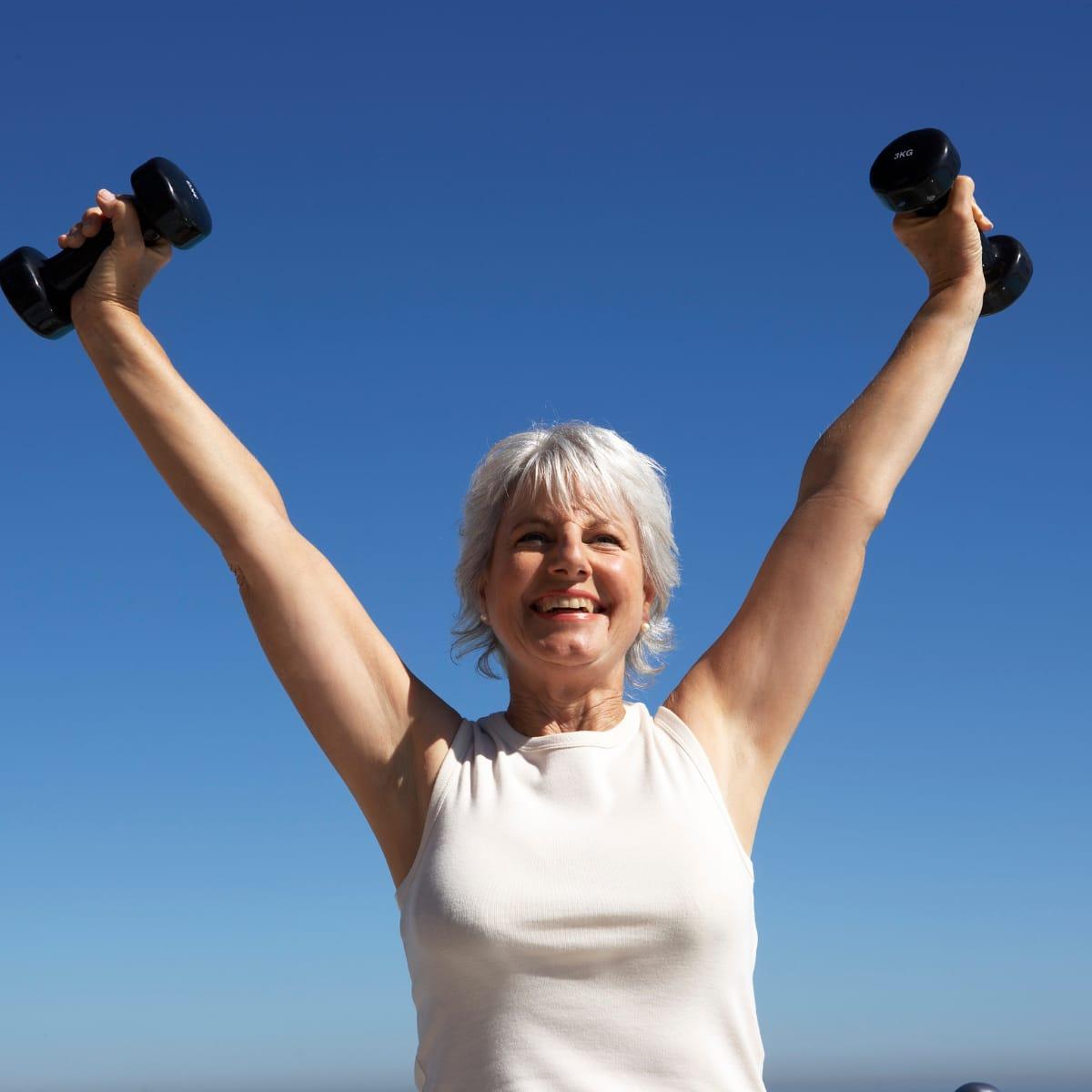 A smiling older woman raising a pair of small dumbbells overhead against a blue sky