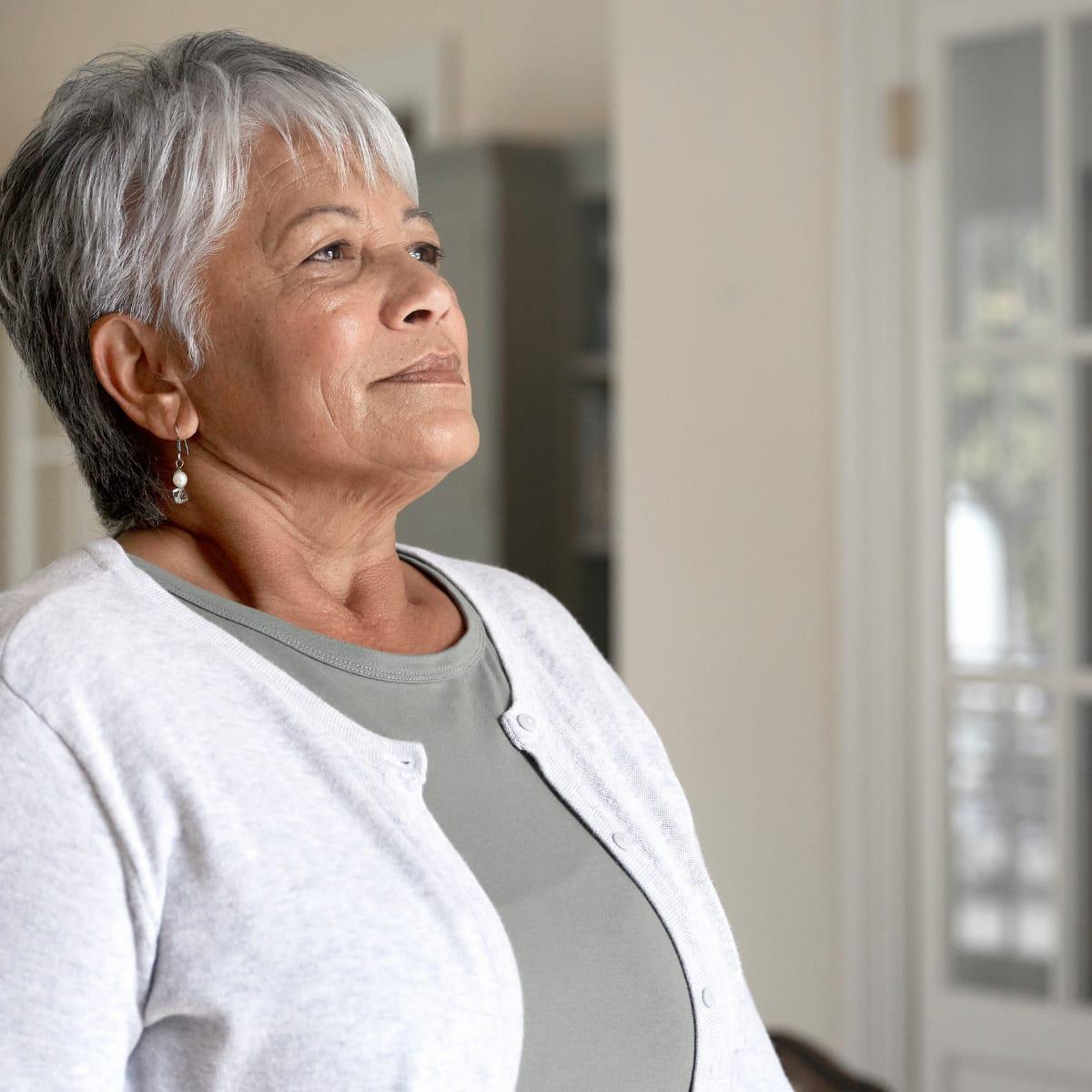 An older woman with short silver hair standing by a window, taking a slow, deep breath