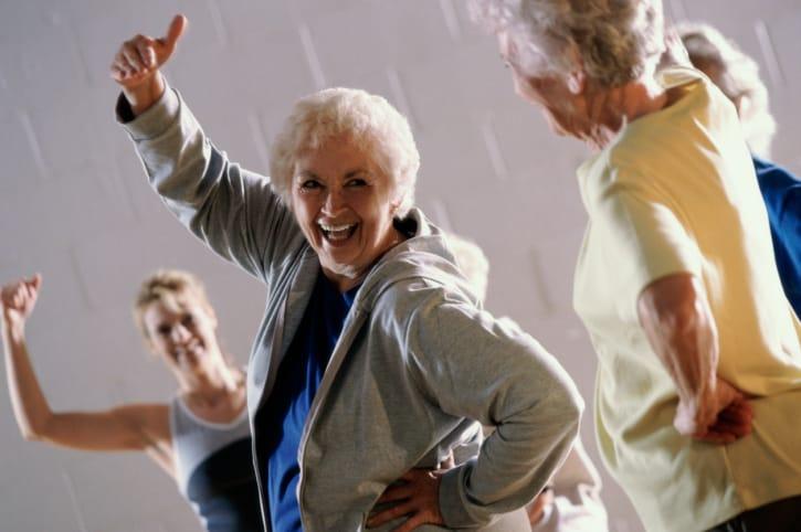 A laughing older woman giving a thumbs-up during a lively group dance class