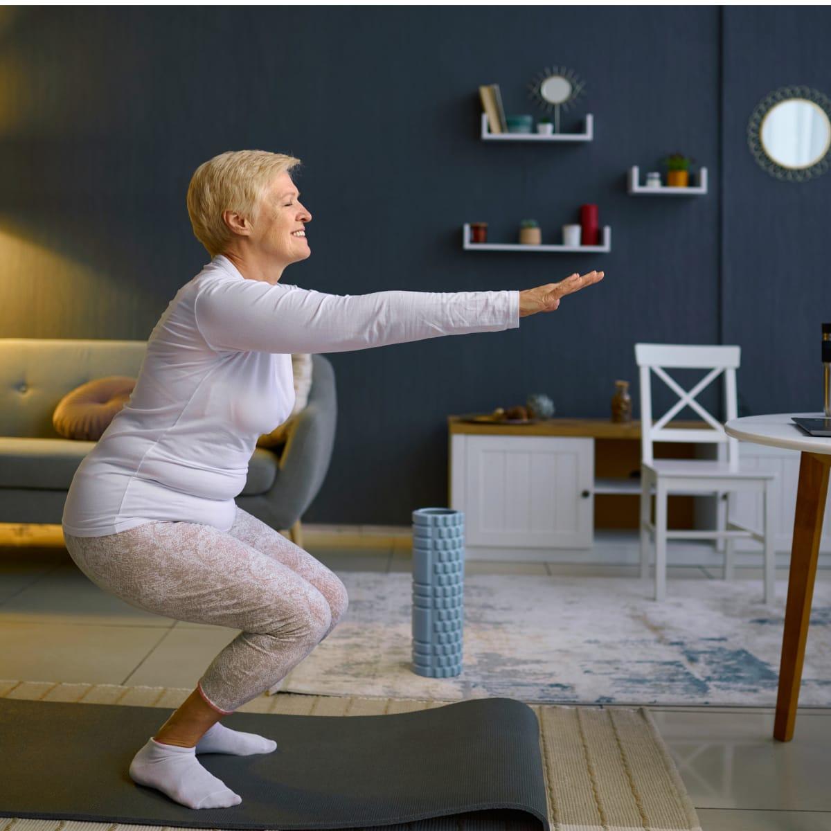 An older woman doing a bodyweight squat on a mat in her living room with her arms reaching forward