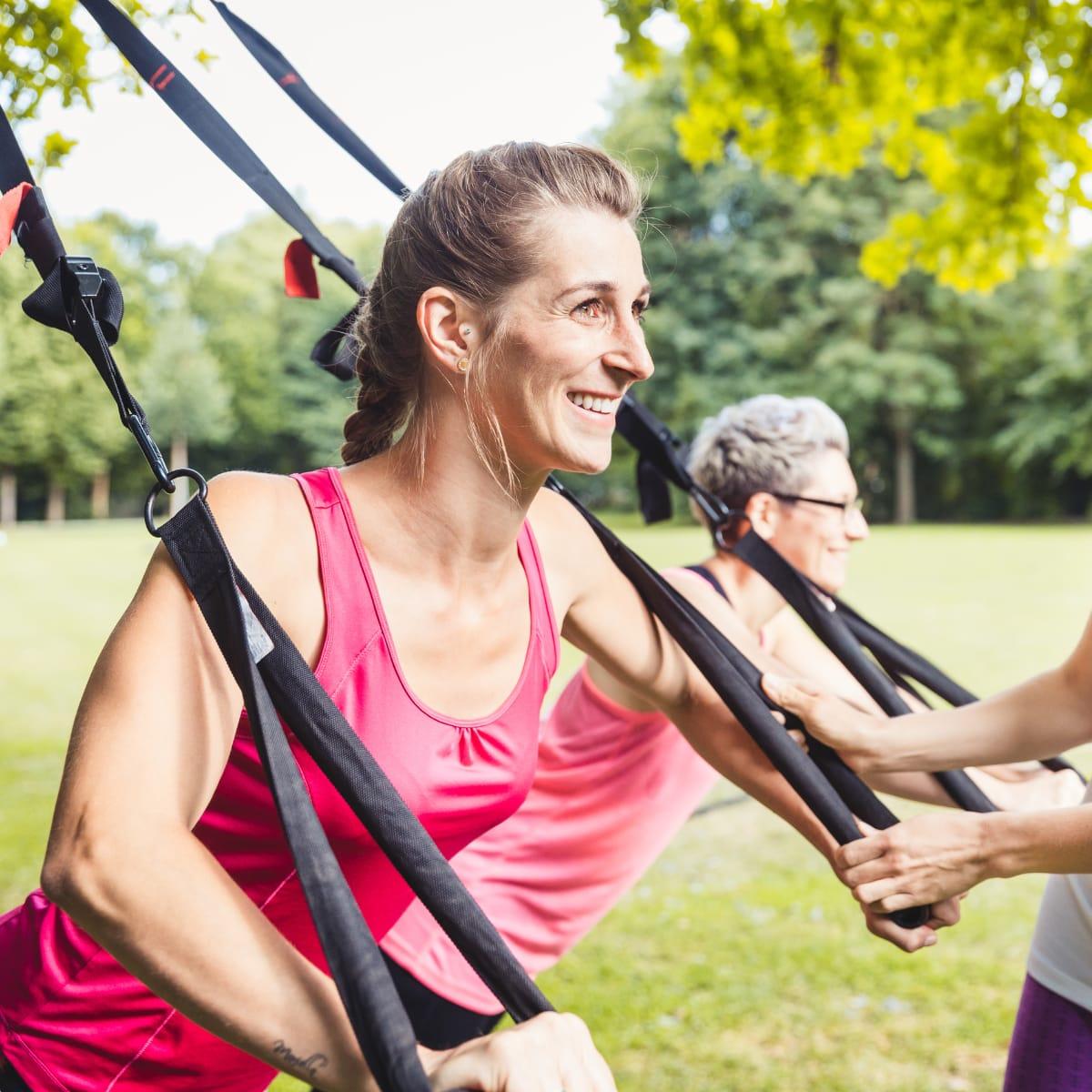 Two women smiling as they lean back holding the handles of suspension training straps outdoors