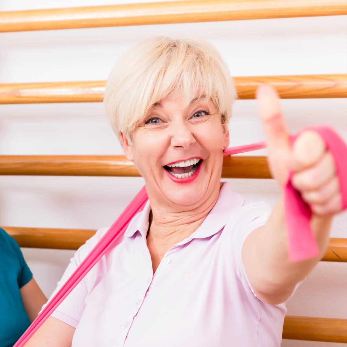A laughing older woman with short blonde hair giving a thumbs-up while holding a pink resistance band