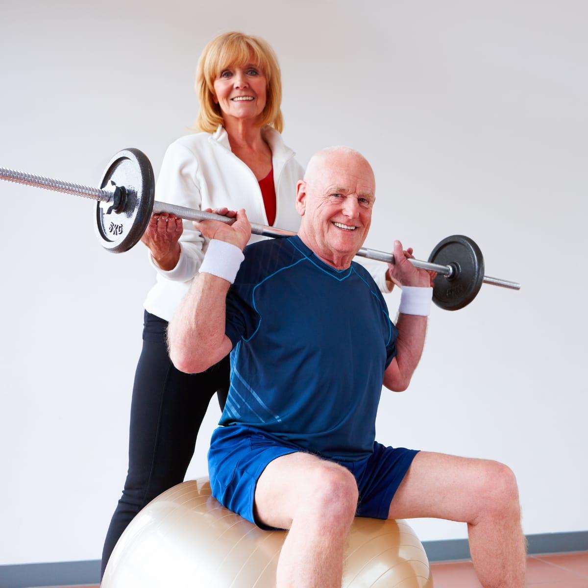 An older man pressing a barbell while seated on an exercise ball, with a smiling woman spotting him