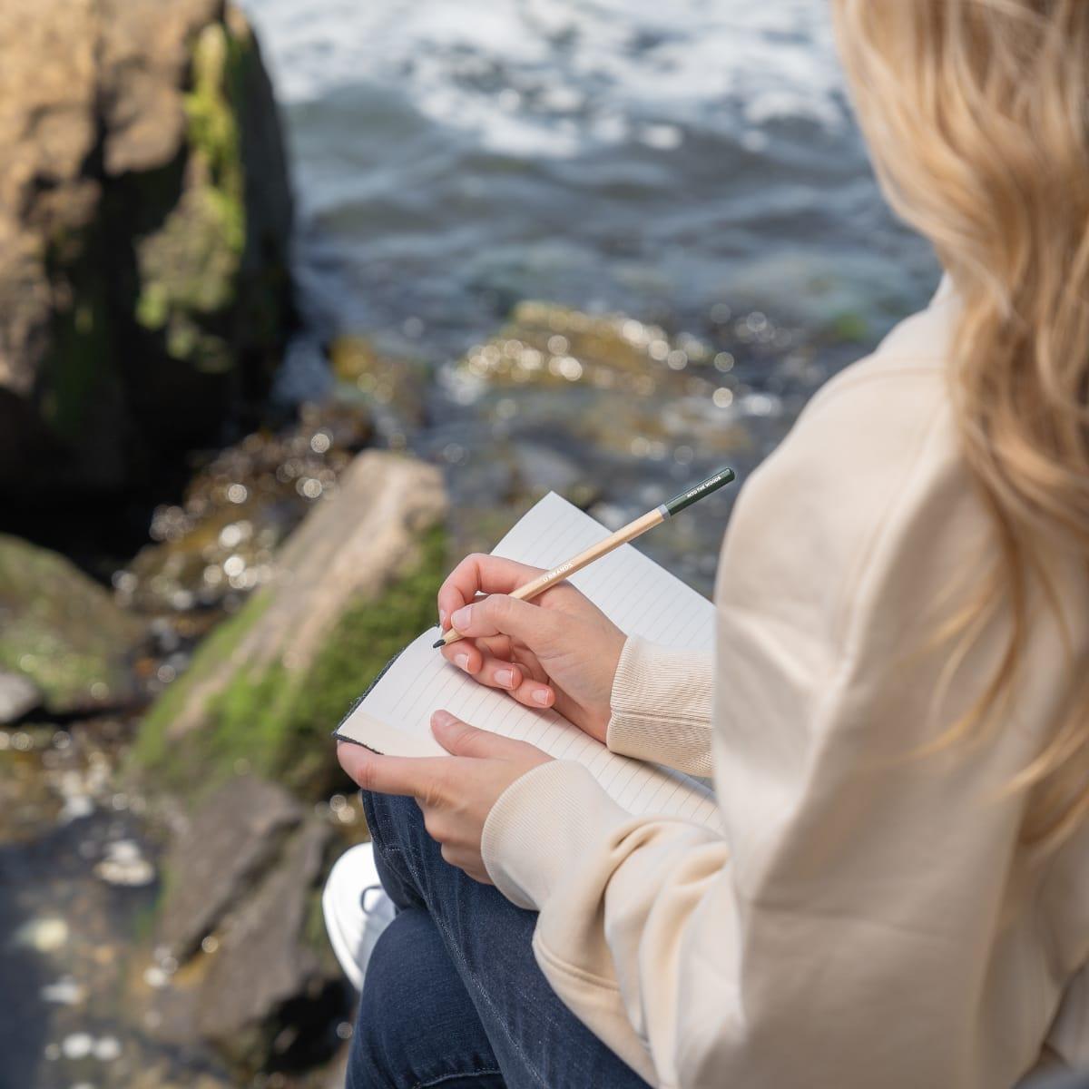 A woman with long blonde hair sitting on rocks by the water, writing in a notebook