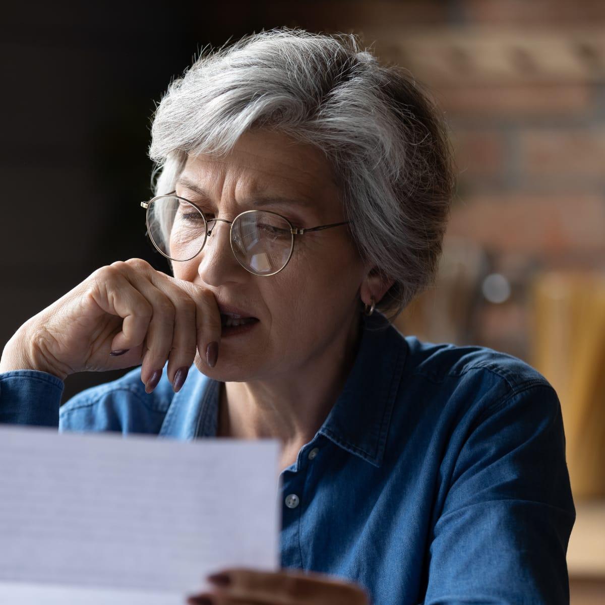 A worried older woman with gray hair and glasses reading a letter, her hand pressed to her mouth