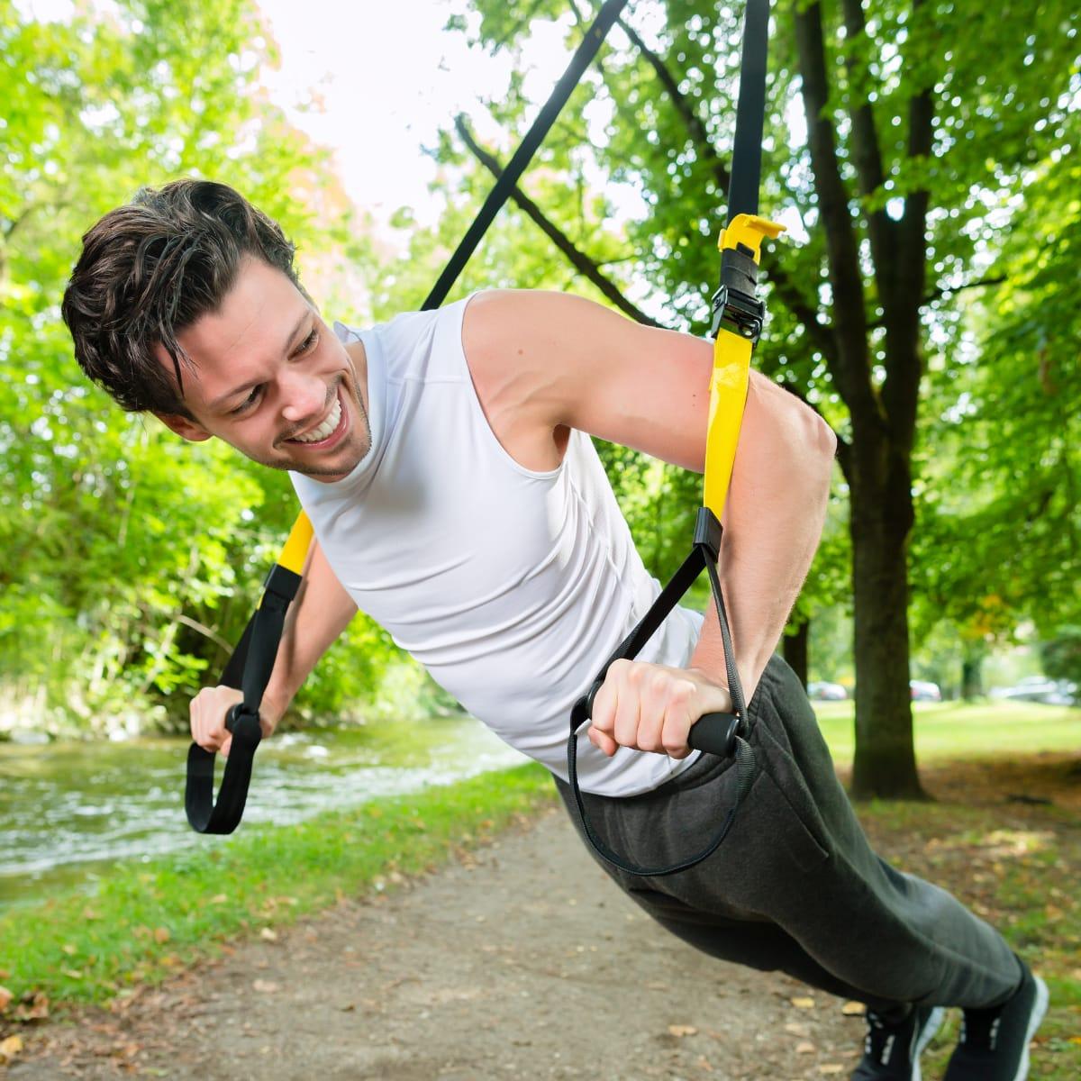 A man leaning back into a suspension trainer strap row under trees in a park