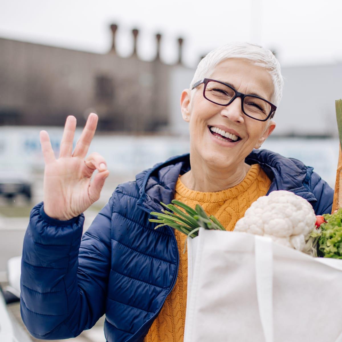 A smiling older woman in glasses giving an OK sign while carrying a bag of fresh vegetables