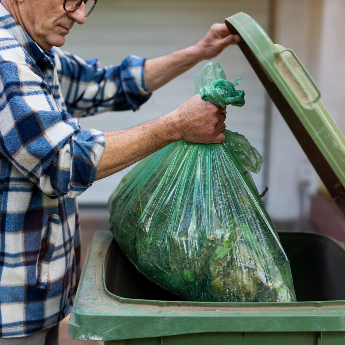 An older man in a plaid shirt lifting a full garbage bag into an outdoor bin with one hand