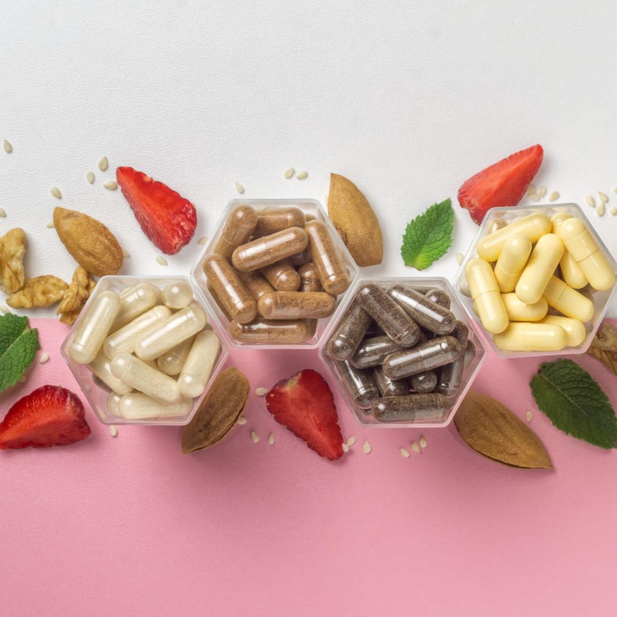 Four hexagonal dishes of different supplement capsules surrounded by almonds, walnuts, strawberries and mint leaves