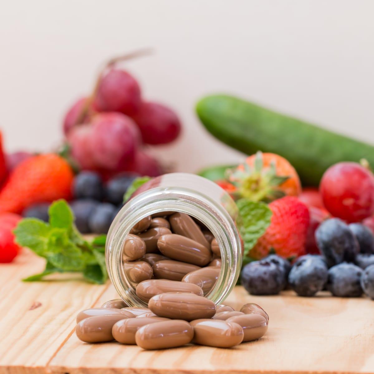 A tipped-over glass jar of brown softgel capsules in front of fresh berries, grapes and a cucumber