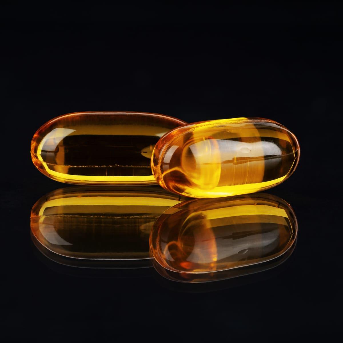 Two golden fish oil softgel capsules reflected on a black surface